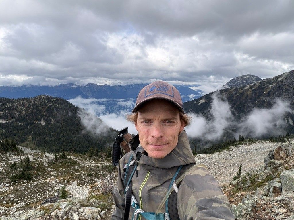 Cody Nicoll on a rocky ridge above the clouds, mountains behind him.